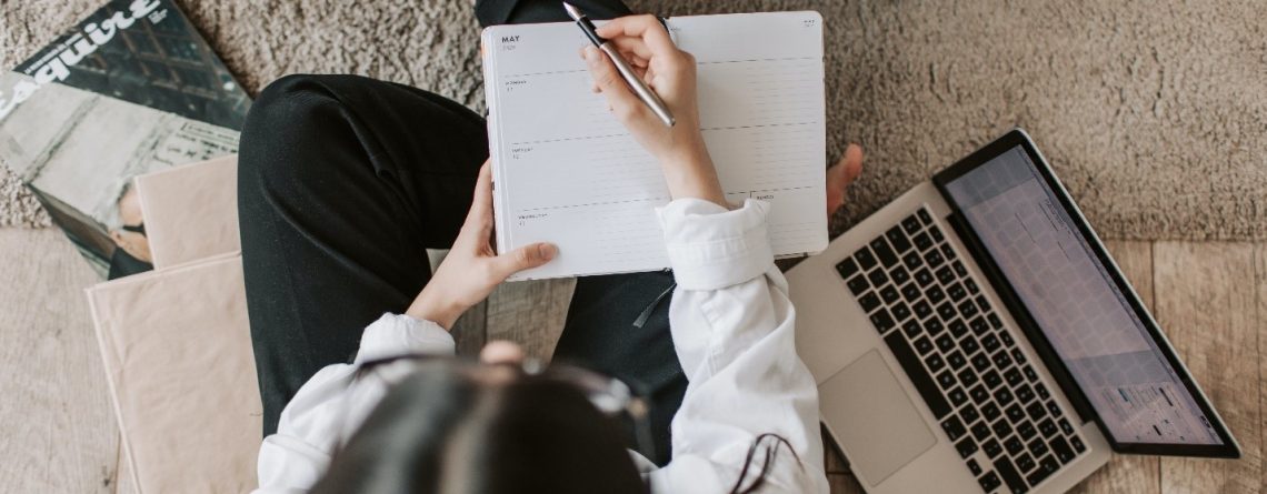 A woman at a desk, writing in a notebook and using a laptop is looking for strategies to start and finish her essay on a perfect note.