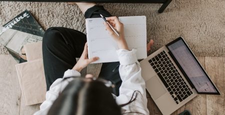 A woman at a desk, writing in a notebook and using a laptop is looking for strategies to start and finish her essay on a perfect note.